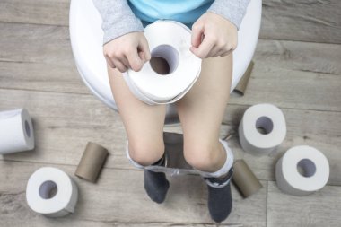 Çocuk beyaz bir tuvalette oturuyor. Elinde tuvalet kağıdı var. Yerde bir sürü rulo var. yakın plan.