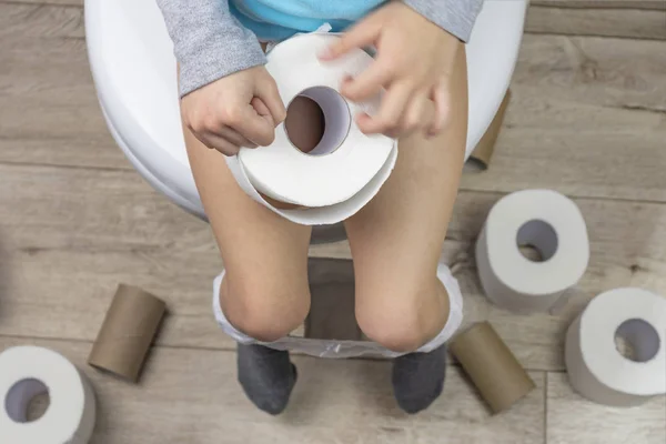 Το παιδί κάθεται σε μια λευκή τουαλέτα. στα χέρια του είναι χαρτί τουαλέτας. Υπάρχουν πολλά ρολά στο πάτωμα. κοντινό πλάνο. — Φωτογραφία Αρχείου