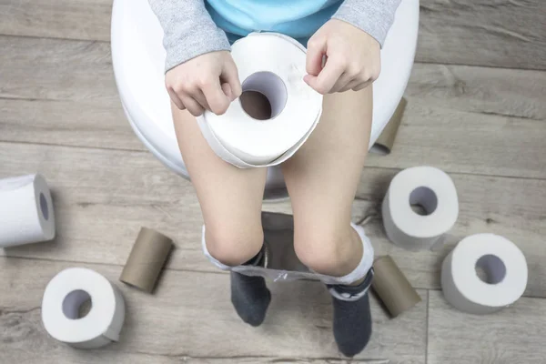 Το παιδί κάθεται σε μια λευκή τουαλέτα. στα χέρια του είναι χαρτί τουαλέτας. Υπάρχουν πολλά ρολά στο πάτωμα. κοντινό πλάνο. — Φωτογραφία Αρχείου