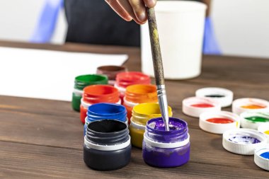 Eski tarz ahşap arka plan. Kutularda boya ve boya fırçası var. Beyaz tişörtlü bir çocuk fırçayı boyaya batırıyor. Yakın plan. Renkli
