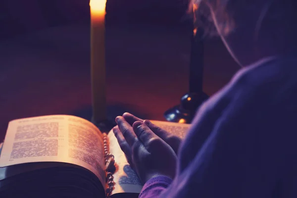 Un cuarto oscuro, una vela está ardiendo. Libro. Sobre la mesa hay una cruz. La niña cruzó los brazos sobre el libro. Hay un tinte . —  Fotos de Stock