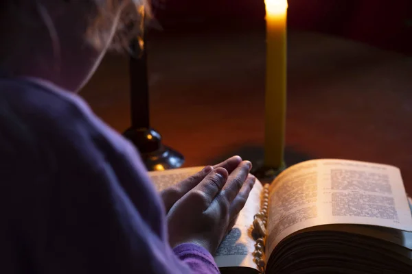 Un cuarto oscuro, una vela está ardiendo. Libro. Sobre la mesa hay una cruz. La niña cruzó los brazos sobre el libro. Hay un tinte . —  Fotos de Stock