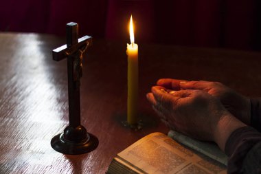 Tahta masa eski usul. Üzerinde bir mum ve bir haç var. Yaşlı bir kadının elleri kitaptan üstündür. Avuçlar katlandı