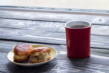 Koyu ahşap arka planda bir doku var. Bir fincan sade kahve ve içinde dolgu malzemesi olan ev yapımı hamur işlerinden iki çörek.
