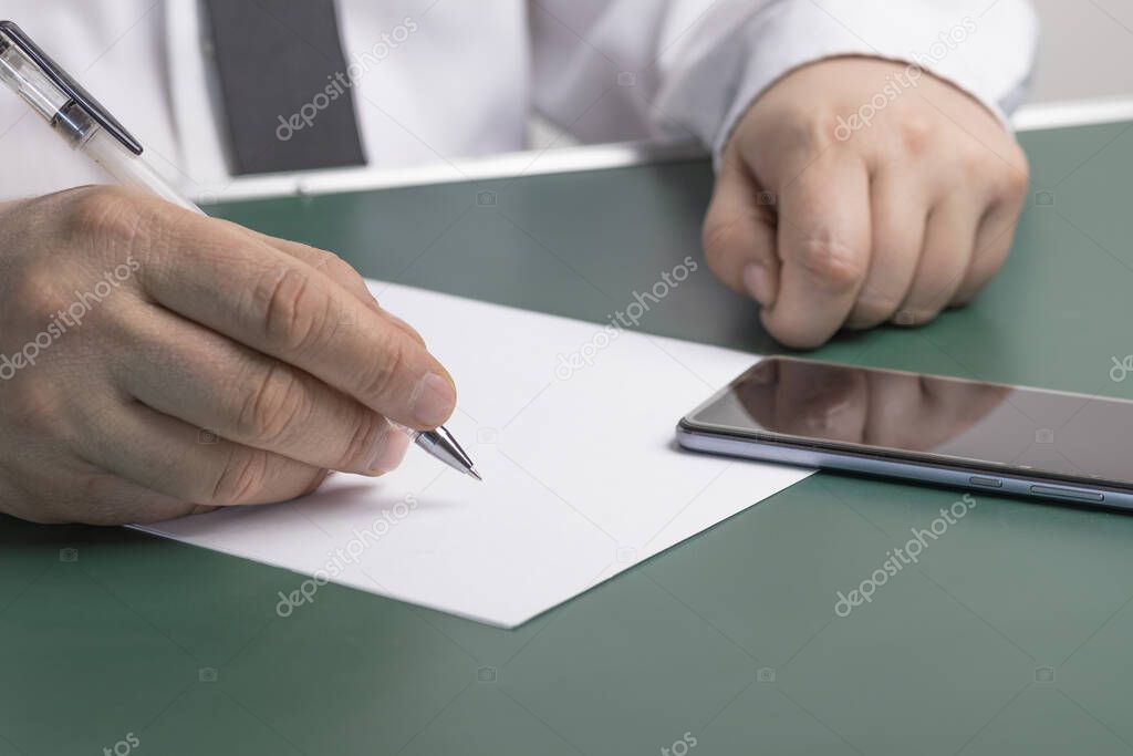 the businessman writes on a sheet of white paper next to the phone. Wood background. Dressed in a shirt and tie. Close-up