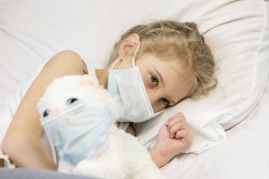 Yüzünde tıbbi maske olan beyaz saçlı bir kız. Yakınlarda, yüzündeki yumuşak oyuncak aynı zamanda bir öksürük maskesi. Yakın plan..