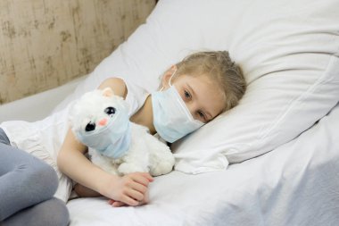Yüzünde tıbbi maske olan beyaz saçlı bir kız. Yakınlarda, yüzündeki yumuşak oyuncak aynı zamanda bir öksürük maskesi. Yakın plan..
