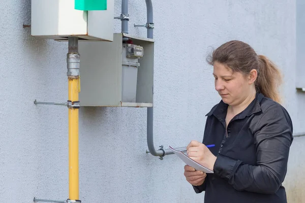 The girl the peasant woman will check on the wall and write down the gas meter in a notebook. Analysis of energy consumption. — Stock Image Girl Peasant Woman Check Wall Write Gas Meter Notebook Analysis Stock Image