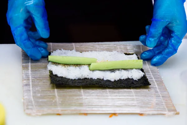 Przygotowanie bułki w sushi bar. Profesjonalny kucharz niebieski w rękawiczkach przygotowuje tradycyjne potrawy japońskie. Ryż, pasztet, ogórek. — Zdjęcie stockowe