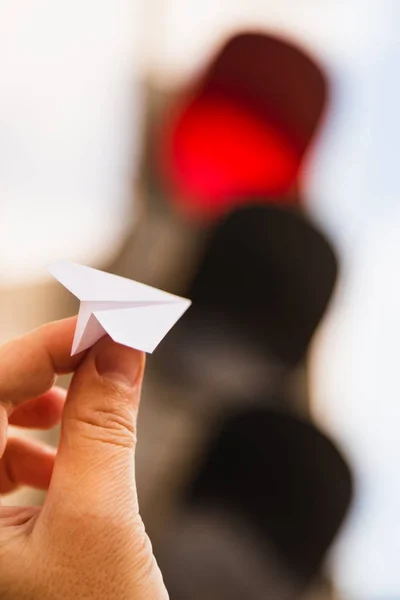 One small paper airplane in hand against a traffic light background. Glowing red prohibition signal. Flight is prohibited. Take-off is impossible, delayed, postponed or canceled. Vertical. Concept. — Stock Photo, Image