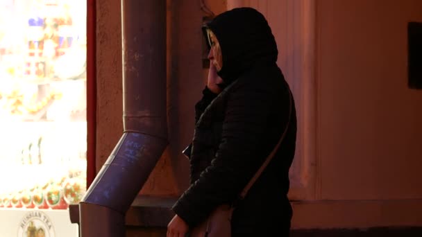 Moscú, Rusia - 26 de octubre de 2019: Una mujer en la calle hablando emocionalmente en un teléfono celular cerca de un escaparate iluminado a finales de otoño por la noche. Señora en una chaqueta caliente con capucha — Vídeos de Stock