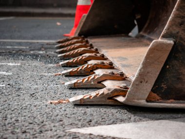 Excavator with hydraulic bucket stands on asphalt surface carriageway. Steel bucket tines close-up. Heavy powerful construction equipment. Emergency red beacon guardrail. Emergency work on sunny day