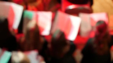 Senfoni orkestrası müzisyenleri sahnede. Koro arkada durup dans ediyor. Bulanık odak. Kemancılar ve viyolonselciler aydınlatılmış müzik standlarının yanındaki sandalyelerde oturuyorlar. Tiyatro ışığındaki renkli noktalar