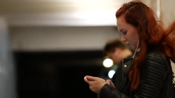 Chica de pelo largo está de pie con un teléfono móvil mientras espera un tren . — Vídeo de stock