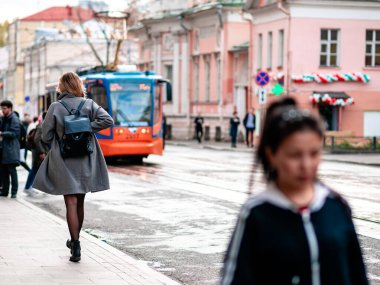 Kısa paltolu, tanınmayan sarışın kadın tramvaya doğru koşuyor. Sırtında siyah bir sırt çantası var. Siyah çoraplı bir kadın tramvay durağına doğru yürüyor. Sıcak, güneşli bir sonbahar günü
