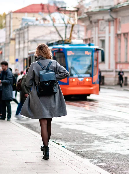 Неузнаваемая молодая блондинка в коротком пальто бросается в трамвай. Черный стильный рюкзак на спине. Женщина в черных чулках идет по улице к трамвайной остановке. Теплый солнечный осенний день — стоковое фото