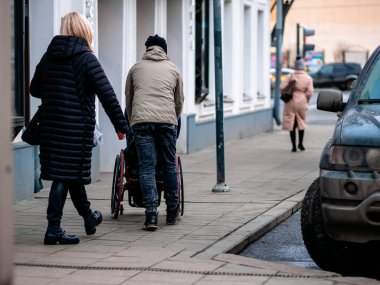 Moskova, Rusya - 17 Ocak 2020: Bir adam kaldırımda fayanslarla döşenmiş tekerlekli sandalyede birini taşıyor. Onlara bir kadın bakıyor. Şehirde park halindeki arabalar..