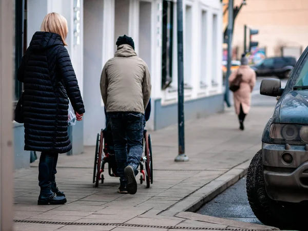 Moscú, Rusia - 17 de enero de 2020: Un hombre lleva a una persona en silla de ruedas a lo largo de la acera pavimentada con azulejos. Una mujer los cuida. Coches aparcados en la ciudad . —  Fotos de Stock