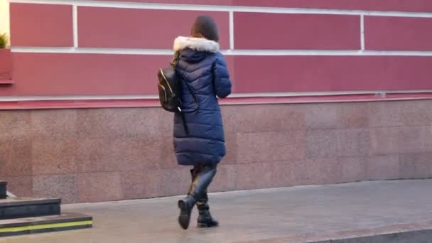 En ung flicka skyndar sig nerför gatan i mörk rock. Kvinna med ryggsäck — Stockvideo