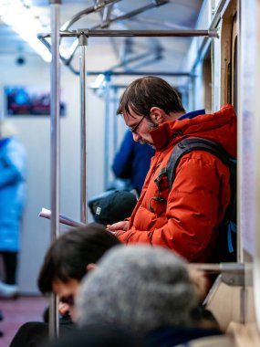 Moskova, Rusya - 8 Şubat 2020: Adam metroya biniyor ve dergi okuyor. Gözlüklü ve turuncu ceketli adam elinde kitapla metro treninin kapısında duruyor..