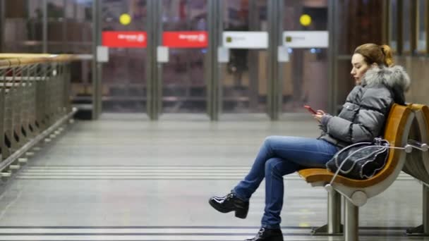 Junge hübsche Dame sitzt auf Holzbank in geräumiger, beleuchteter Lobby mit Telefon — Stockvideo