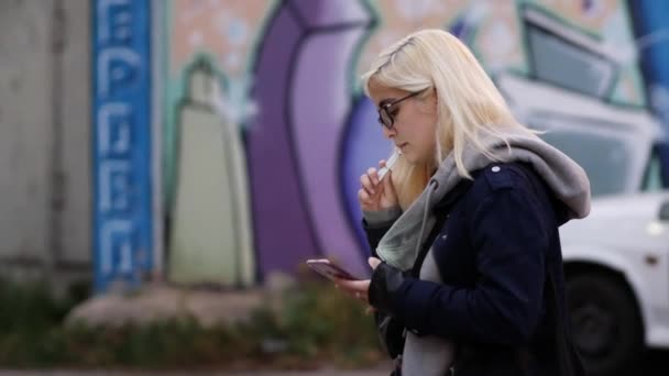 Blond in bril rookt een elektronische sigaret en kijkt naar smartphone scherm — Stockvideo