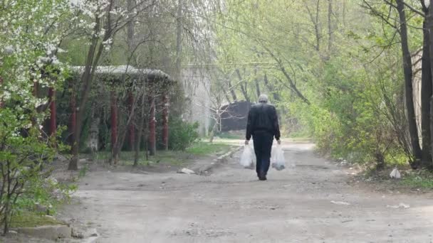 Un homme âgé portant deux sacs lourds avec des provisions dans la rue de la ville rentre chez lui à pied. — Video