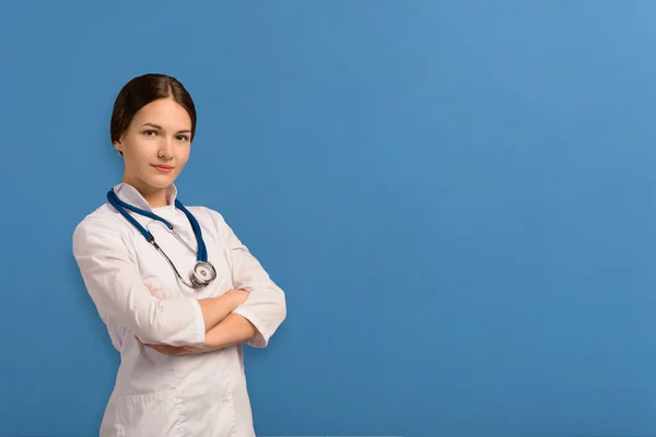 Gülümseyen genç bir doktorun portresi. Beyaz bir bornoz giymişti. Mavi bir arka planda çapraz ellerle durarak.. — Stok fotoğraf