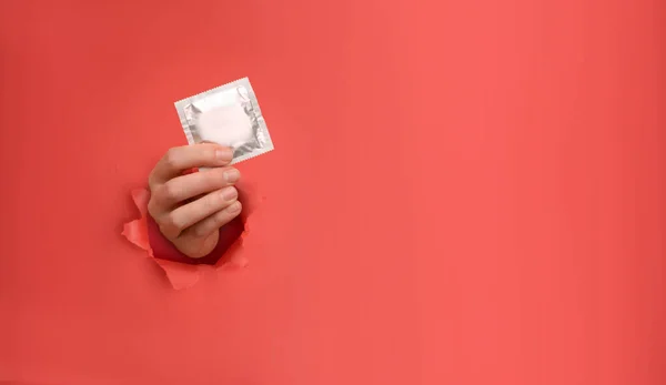 Female hand holds condom on torn red paper wall. Safe pleasure and protection, contraception, protection from AIDS and day to fight AIDS. Copy space aside for your advertising content — Stock Photo, Image