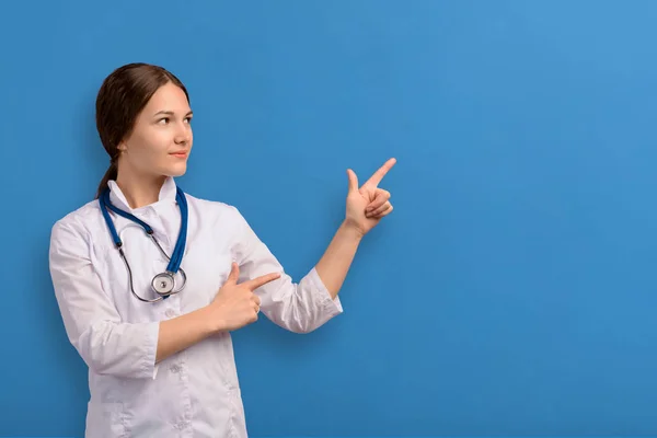 Gülümseyen mutlu doktor uzun saçlı genç kız mavi arka planı işaret ediyor tasarım ve metin için yeri var.. — Stok fotoğraf