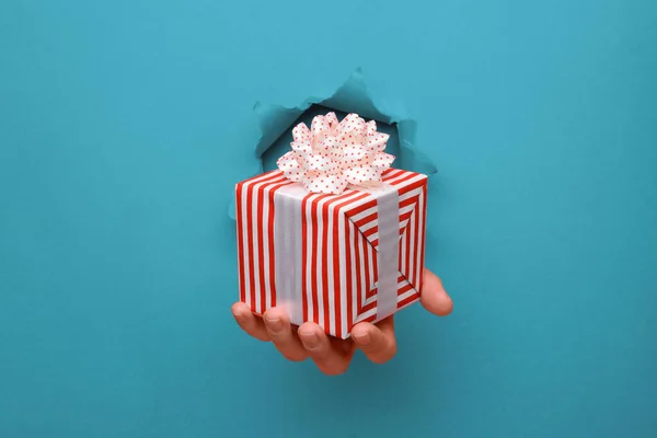 Main donner boîte cadeau rayé rouge et blanc sur le mur de papier bleu déchiré. Espace de copie réservé à votre publicité et à votre contenu d'offre ou de vente . — Photo