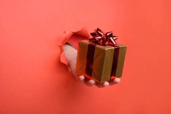 Χέρι δώσει χρυσό κουτί δώρου με κόκκινη κορδέλα σε σχισμένο κόκκινο τοίχο από χαρτί. Αντιγραφή χώρο στην άκρη για τη διαφήμισή σας και την προσφορά ή την πώληση περιεχομένου. — Φωτογραφία Αρχείου