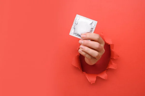 Hand give condom on torn red paper wall. Safe pleasure and protection, contraception, protection from AIDS and day to fight AIDS. Copy space aside for your advertising content — Stock Photo, Image