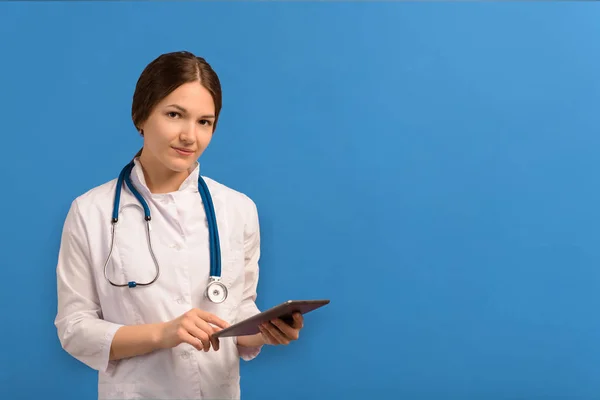 Beyaz önlüklü, steteskoplu, mavi arka planda tablet tutan güzel bir kadın doktor.. — Stok fotoğraf