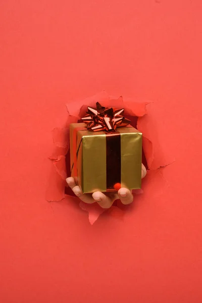 Hand geben Gold-Geschenk-Box mit rotem Band auf zerrissenen roten Papierwand. Kopierfläche beiseite für Ihre Werbung und Angebots- oder Verkaufsinhalte. — Stockfoto