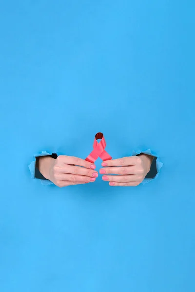 Red ribbon is symbol of fight against AIDS or disease cancer of female breast. Girl's hand holds a red ribbon on a blue torn papper background. Copy space aside for your advertising content — ストック写真