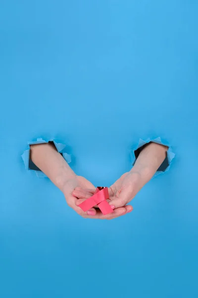 Red ribbon is symbol of fight against AIDS or disease cancer of female breast. Girl's hand holds a red ribbon on a blue torn papper background. Copy space aside for your advertising content — ストック写真