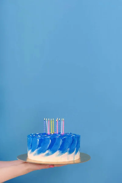 Girl's hands are holding beautiful and tasty cake with white and true blue color cream with holiday candles on a blue background with place for text. — Stock Photo, Image