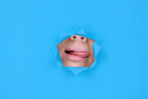 Part of woman\'s face lips and tongue an attractive woman licks her upper lip with her tongue behind broken wall or torn paper, on blue background. Place for text, banner blank and or Feminism design