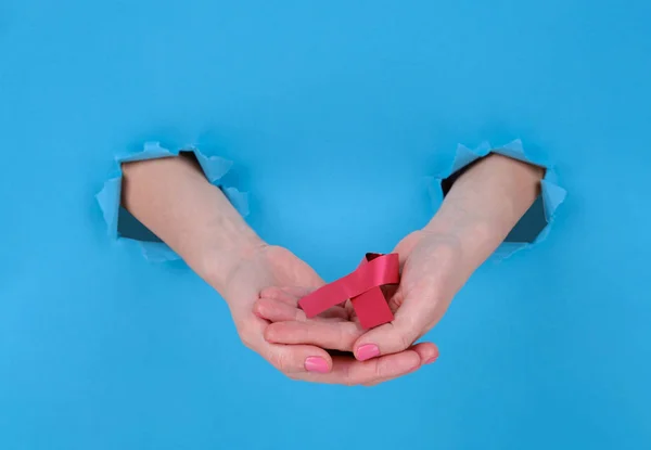 Red ribbon is symbol of fight against AIDS or disease cancer of female breast. Girl's hand holds a red ribbon on a blue torn papper background. Copy space aside for your advertising content — ストック写真