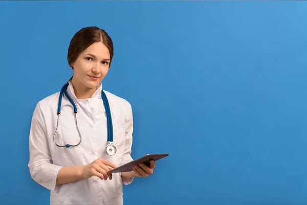 Beyaz önlüklü, steteskoplu, mavi arka planda tablet tutan güzel bir kadın doktor.. — Stok fotoğraf