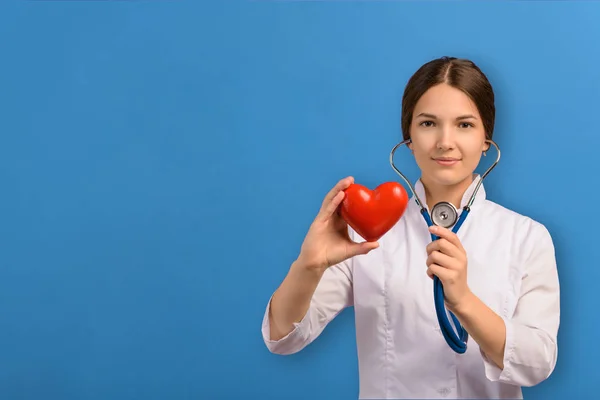 Steteskoplu, beyaz önlüklü, kırmızı kalbi mavi arka planda inceleyen genç bir doktor. Tıp ve kardiyoloji konularının yerleşimi. — Stok fotoğraf