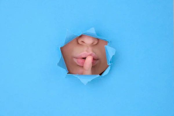 Part female face lips and tongue of attractive woman, girl covers her lips with her finger symbolizes silence behind broken wall or torn paper, blue background. Space for text, feminist banner