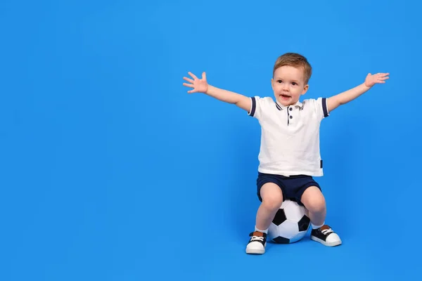 Boy sits on soccer ball and rejoices in victory, raising his arms to top against blue background. Emotions of winner and champion, place for text. Concept of winning and achieving goal. — Stock Photo, Image