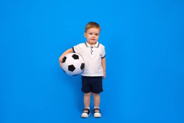 Çocuk mavi arka planda bir futbol topuyla duruyor..