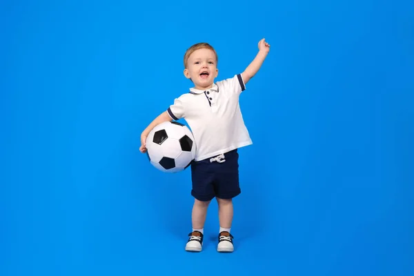 Mutlu gülümseyen çocuk futbol topuyla ayakta duruyor ve zaferle seviniyor, kollarını mavi arka plana doğru kaldırıyor. Kazananın ve şampiyonun duyguları, metne dökülsün. Kazanma ve hedefe ulaşma kavramı