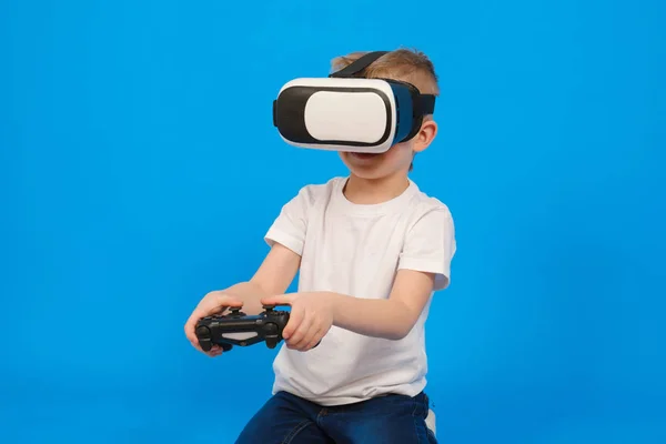 Хлопчик в окулярах VR грає на ігровій шафі, взаємодіючи з віртуальним світом, одягнений у біле поло та джинсові штани на синьому фоні. Інноваційні технології та концепція освіти та розваг . — стокове фото