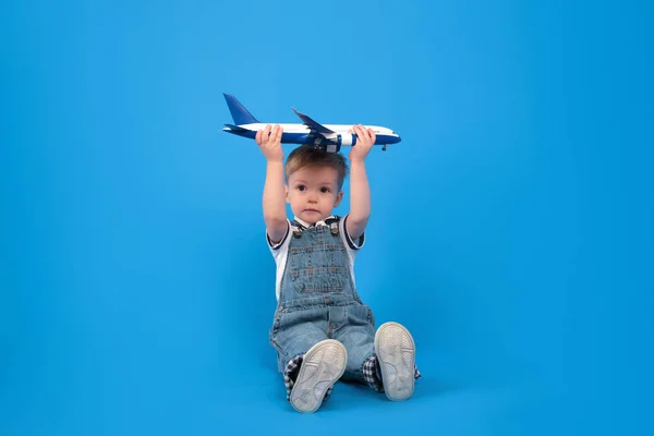 Happy child sits holding plane in his hand and imagining how he is flying on plane on blue background. Imagination, creativity and concept ideas. — Stock Photo, Image