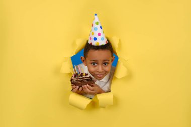 Mutlu Afro-Amerikalı çocuk doğum gününü elinde mumlarla çikolatalı pasta tutarak ve sarı yırtık kağıtta tatilin tadını çıkararak kutluyor. Konsept parti doğum günü, kopya için boşluk