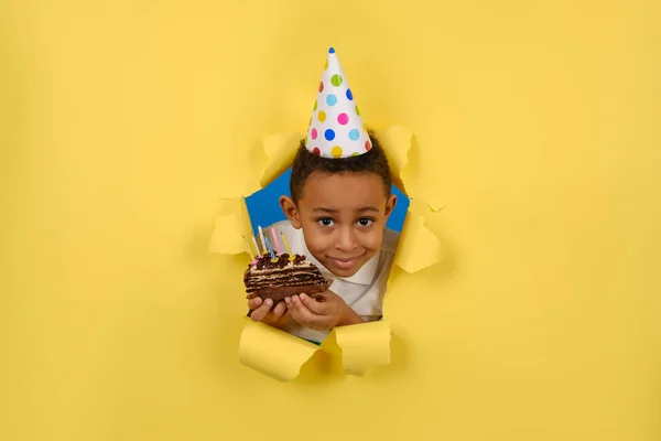 Feliz niño afroamericano celebra su cumpleaños sosteniendo un pastel de chocolate en sus manos con velas y disfrutando de las vacaciones en un fondo de papel amarillo roto. Concepto fiesta cumpleaños, espacio para la copia — Foto de Stock